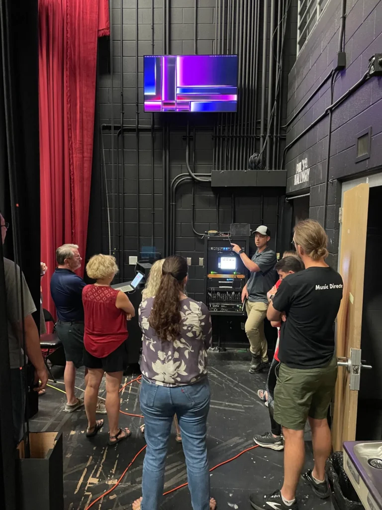People gathered backstage near technical equipment and a screen.
