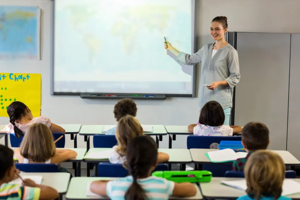 Teacher pointing at a world map in a classroom while students listen.