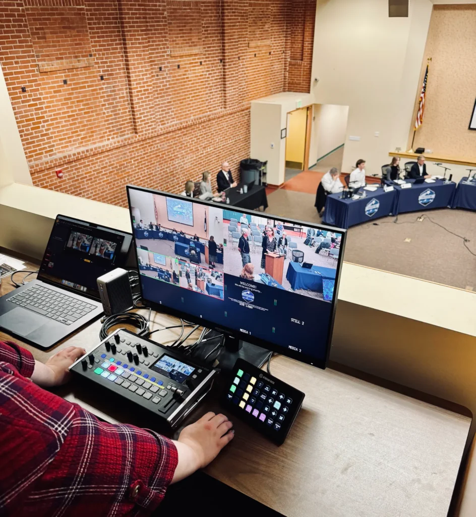Person operating video equipment overlooking a meeting room with multiple monitors and controls.