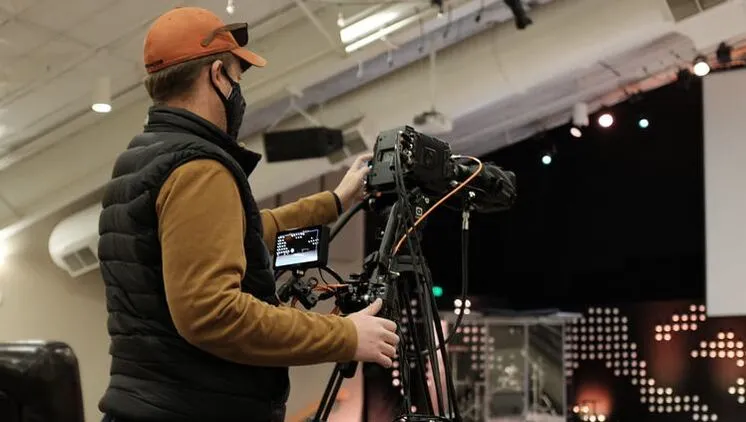 Person operating a professional video camera on a tripod in an indoor setting.
