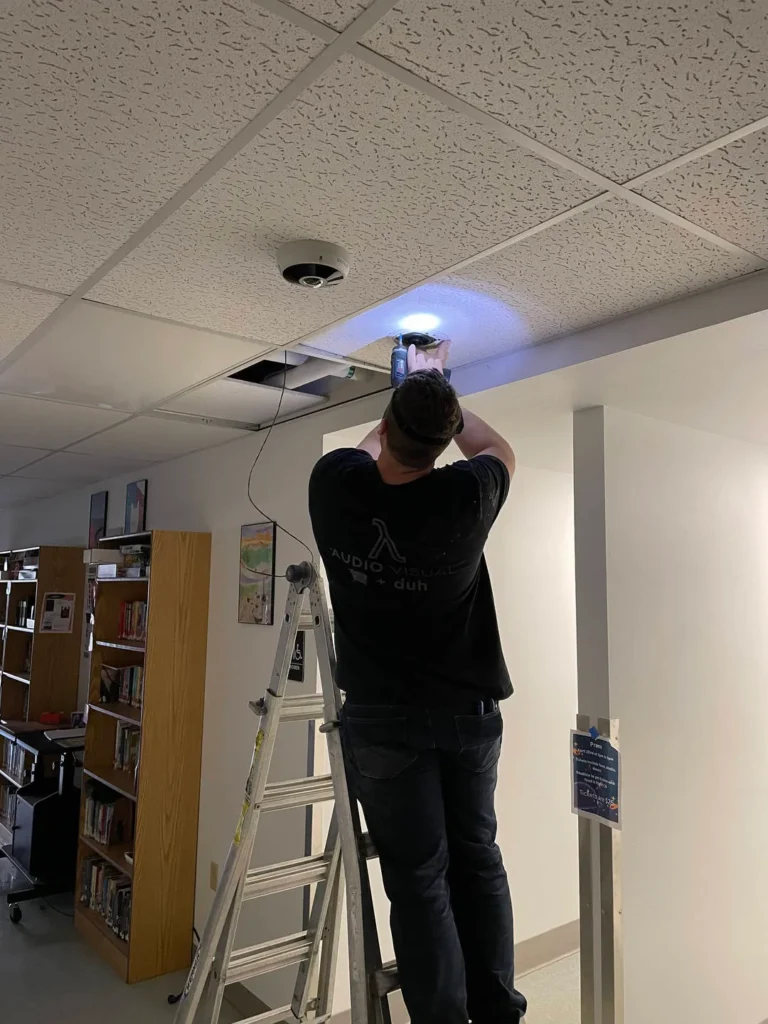 A person on a ladder works on a ceiling panel with a power tool.