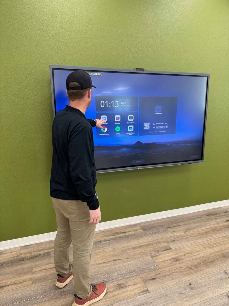 Person pointing at a large touchscreen display in a room with green walls.
