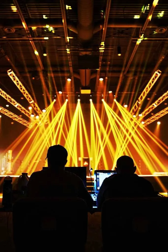 Silhouettes of two people operating equipment at a concert with bright stage lights.